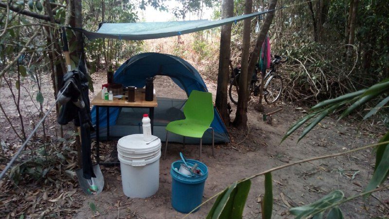 A dome tent under a green tarp, with a small table, a white bucket, a blue bucket and a green chair in front