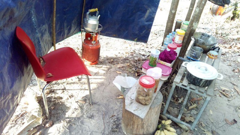 A gas burner with a kettle, a red chair and food containers on a small table, behind a blue tarp
