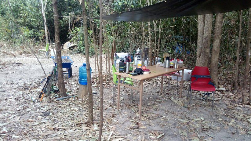 A long table under trees with water bottles, flasks and cups, and a red chair beside it
