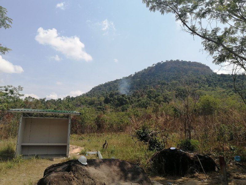 A small open shelter in dry grass, with a forested hill and a thin line of smoke behind it