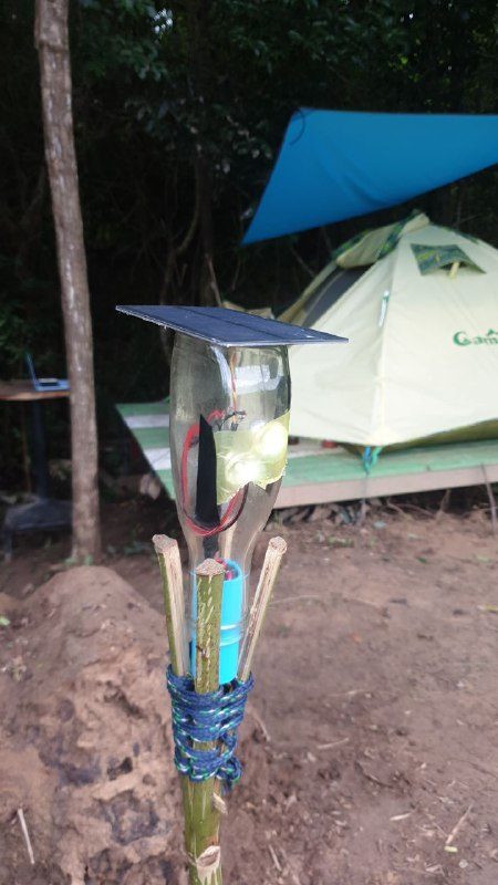 A plastic bottle lamp with a small solar panel on top, tied to a bamboo stick in front of a tent