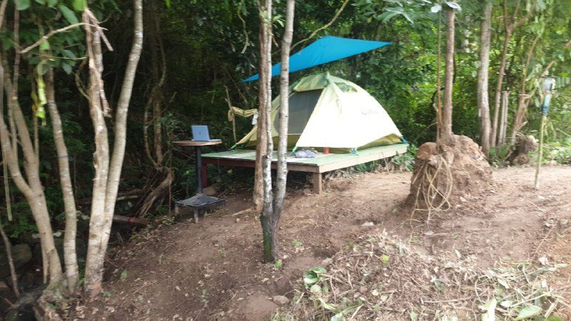 A tent on a raised wooden platform under a blue tarp, with a small table and chair beside it